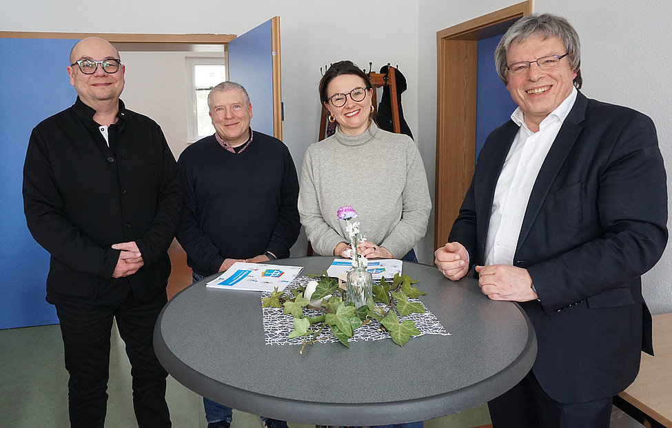 Vier Personen stehen lächelnd um einen runden Tisch mit Blumen und Prospekten, freundliche Stimmung in einem hellen Raum.