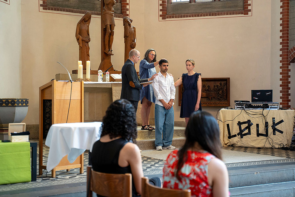 Ein Mann steht zusammen mit drei Personen vor dem Altar einer Kirche, während zwei Zuschauerinnen die Szene aufmerksam aus den vorderen Reihen verfolgen.