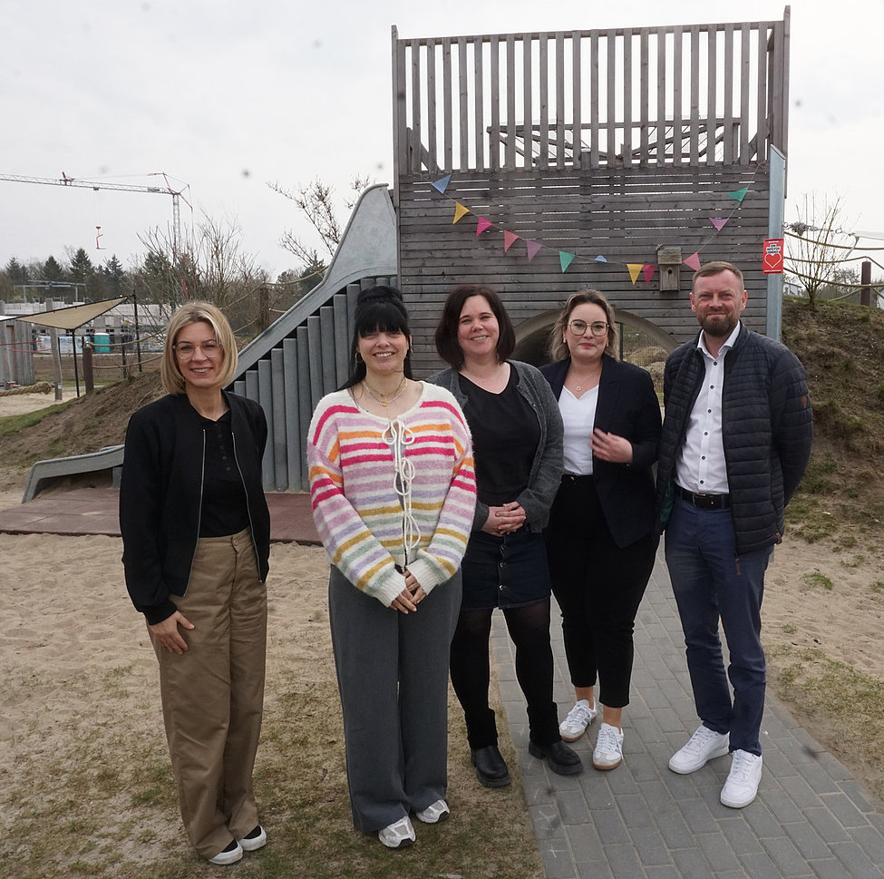 Das Bild zeigt vier Frauen und einen Mann, die in die Kamera lächeln. Im Hintergrund ist ein Kletterturm für Kinder zu sehen.