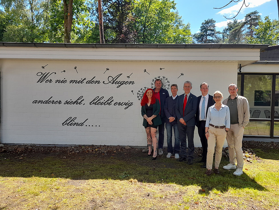Sieben Personen posieren vor einer Wand mit Schriftzug „Wer nie mit den Augen anderer sieht…“ in einem sonnigen Außenbereich, lächelnd.