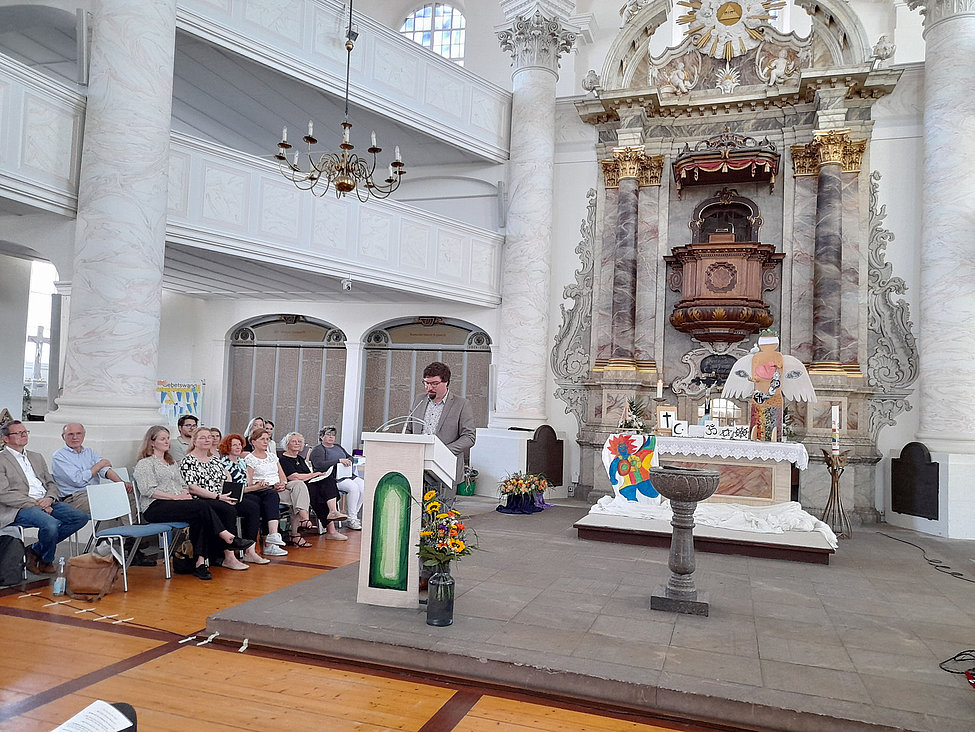Redner am Pult vor Publikum und buntem Engelaltar in Kirche