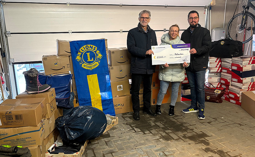 Drei Personen übergeben einen Spendenscheck über 1.000 Euro, umgeben von Hilfspaketen im Lager mit Lions Club Fahne im Hintergrund.