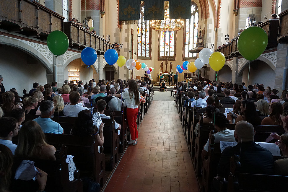 Kircheninnenraum mit gefüllten Bänken, bunter Ballonreihe entlang des Mittelgangs und einer Zeremonie auf der Bühne.