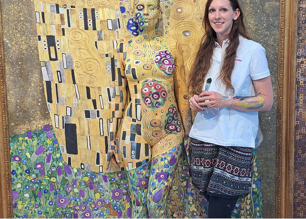 Frau steht lächelnd neben einer lebensgroßen Figur im Gustav‑Klimt‑Stil mit goldenen Mustern; sie hält ein Gerät, Körperbemalung sichtbar.