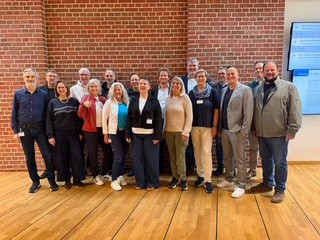 Gruppe von Erwachsenen posiert lächelnd in einem Saal vor roter Ziegelwand – formelles Gruppenfoto in Reihen.