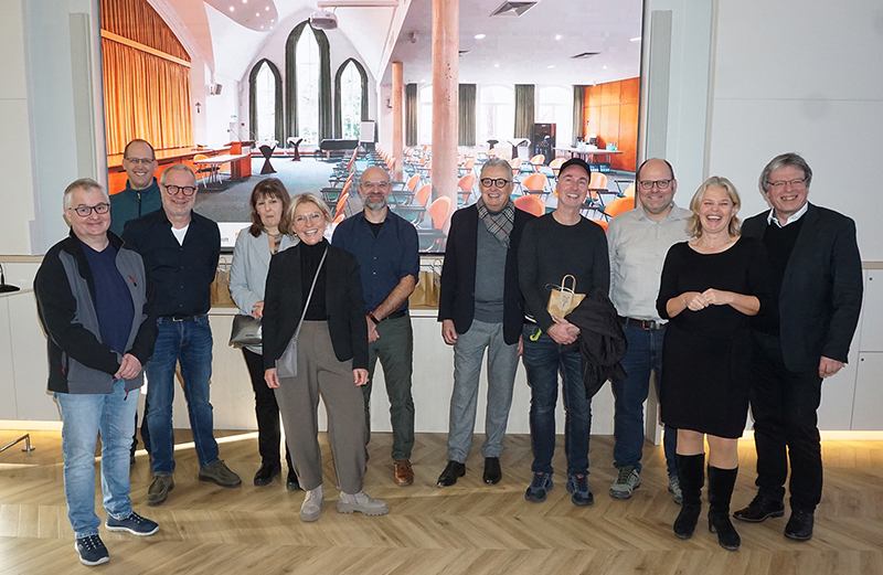 Elf Erwachsene stehen lächelnd in einem Raum vor einem großen Bild mit Saal und Fenstern. Die Gruppe wirkt entspannt und freundschaftlich.